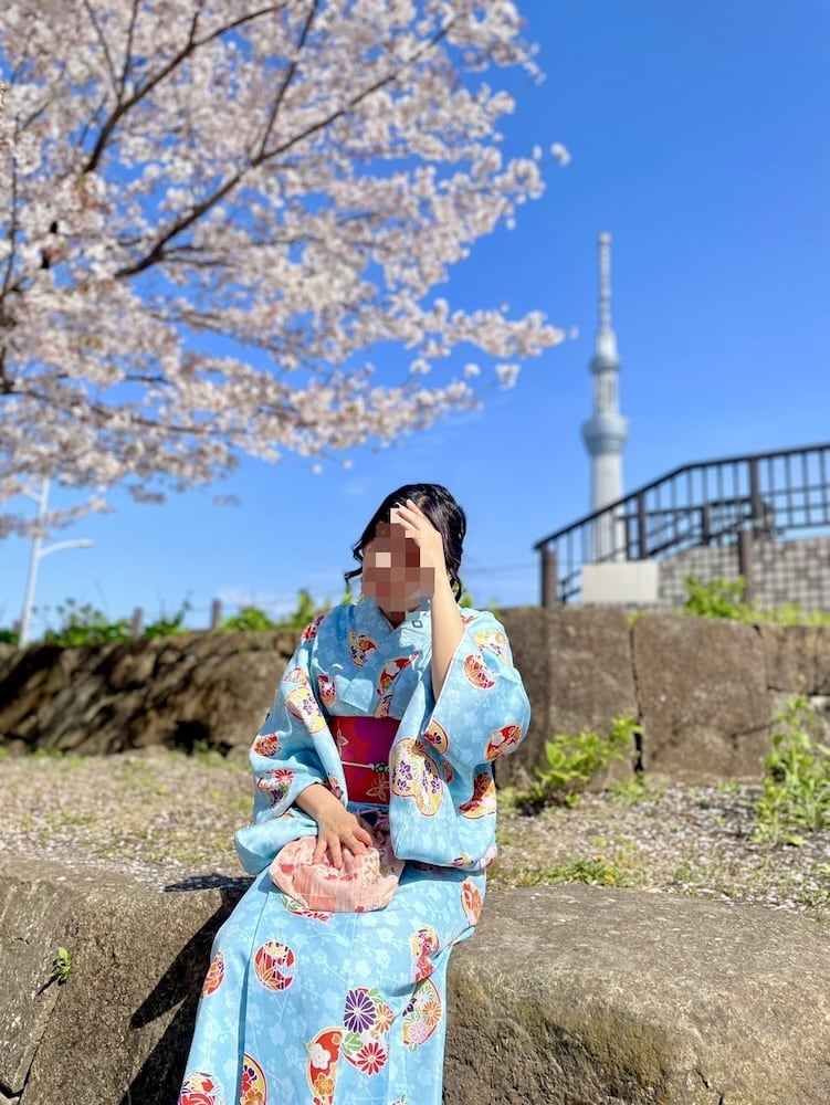 A woman in a Japanese kimono sits on a stone wall, surrounded by cherry blossoms.