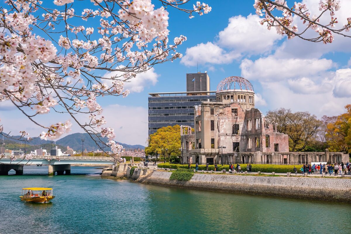 Cherry blossoms frame the Hiroshima Peace Memorial on a sunny day, with a river and boat in the foreground and people walking nearby.