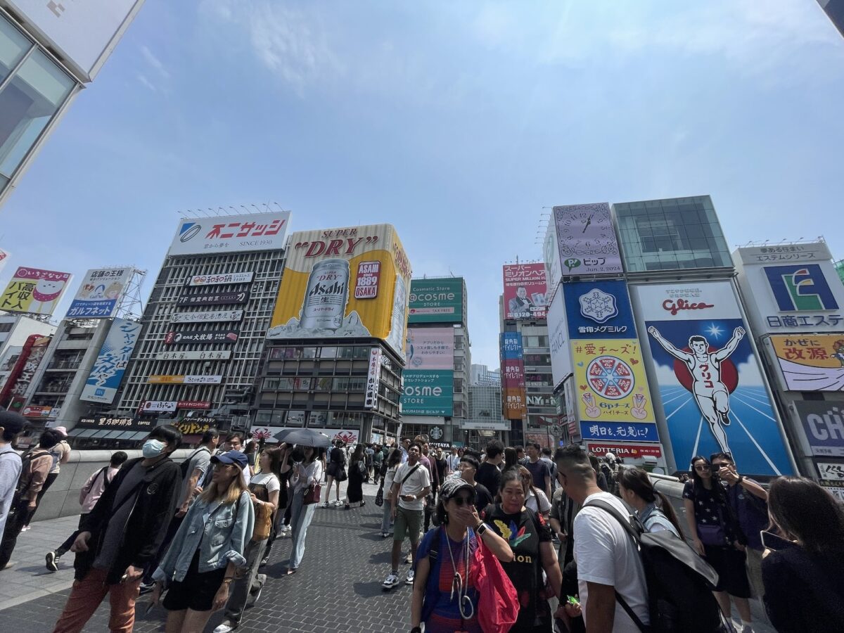 A busy street scene in a city with large colorful billboards, crowds of people walking, and clear blue sky overhead.