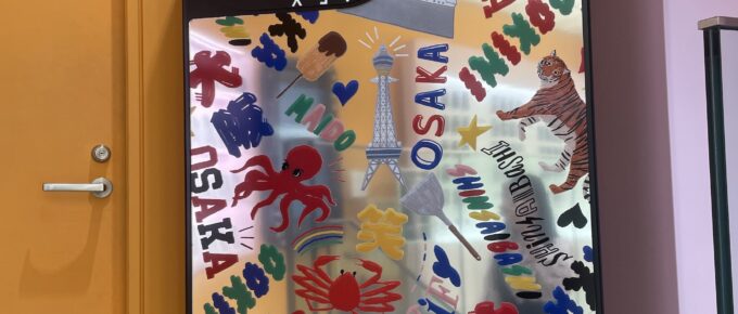A large decorative display shaped like a smartphone features colorful graphics, including the word “OSAKA,” tigers, lions, trees, and a tower illustration, positioned next to a yellow door.