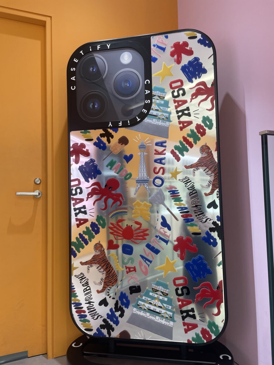A large decorative display shaped like a smartphone features colorful graphics, including the word “OSAKA,” tigers, lions, trees, and a tower illustration, positioned next to a yellow door.