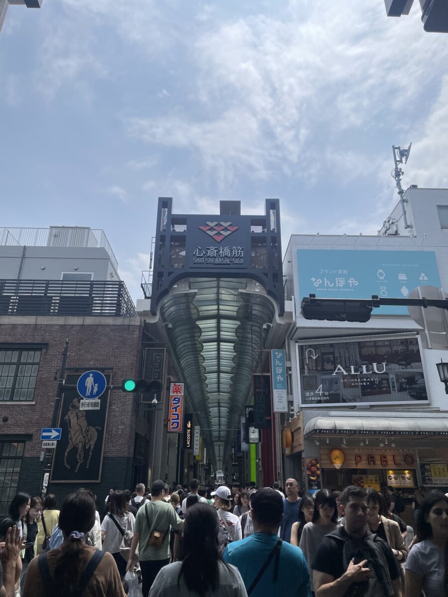 Crowds of people walk through the entrance of a covered shopping street in Shinsaibashi, Osaka, Japan on a sunny day.