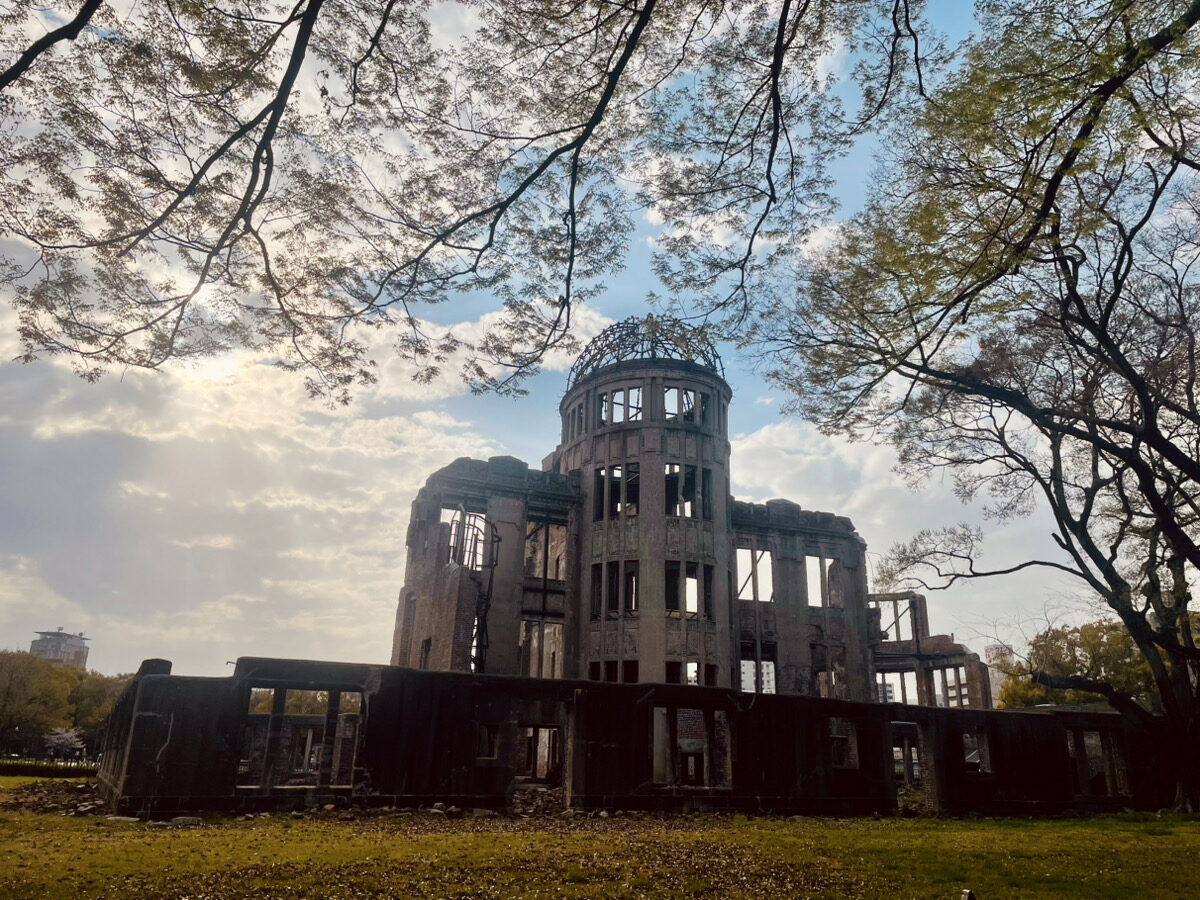 Ruins of the Hiroshima Peace Memorial (Genbaku Dome) stand under a partly cloudy sky, framed by tree branches and surrounded by grass.