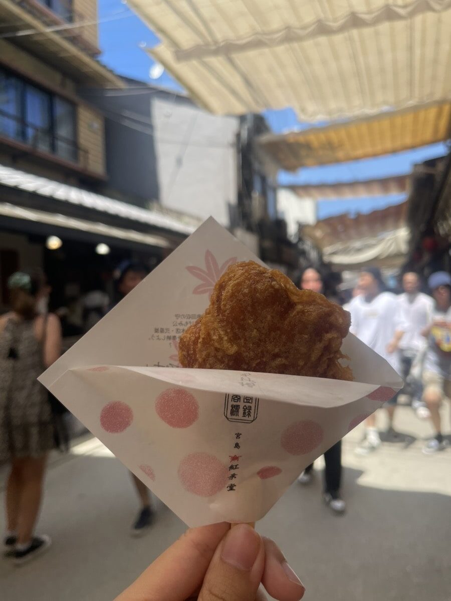 A hand holds a piece of fried food wrapped in white polka-dotted paper on a sunny street with blurred people and buildings in the background.