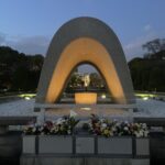 A white stone arch is shown at dusk, with flowers placed in front and the Eternal Flame visible under the arch.