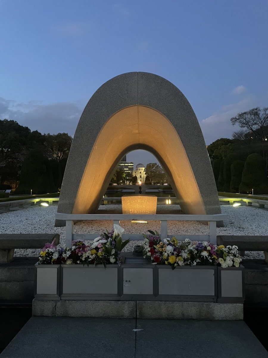 A white stone arch is shown at dusk, with flowers placed in front and the Eternal Flame visible under the arch.