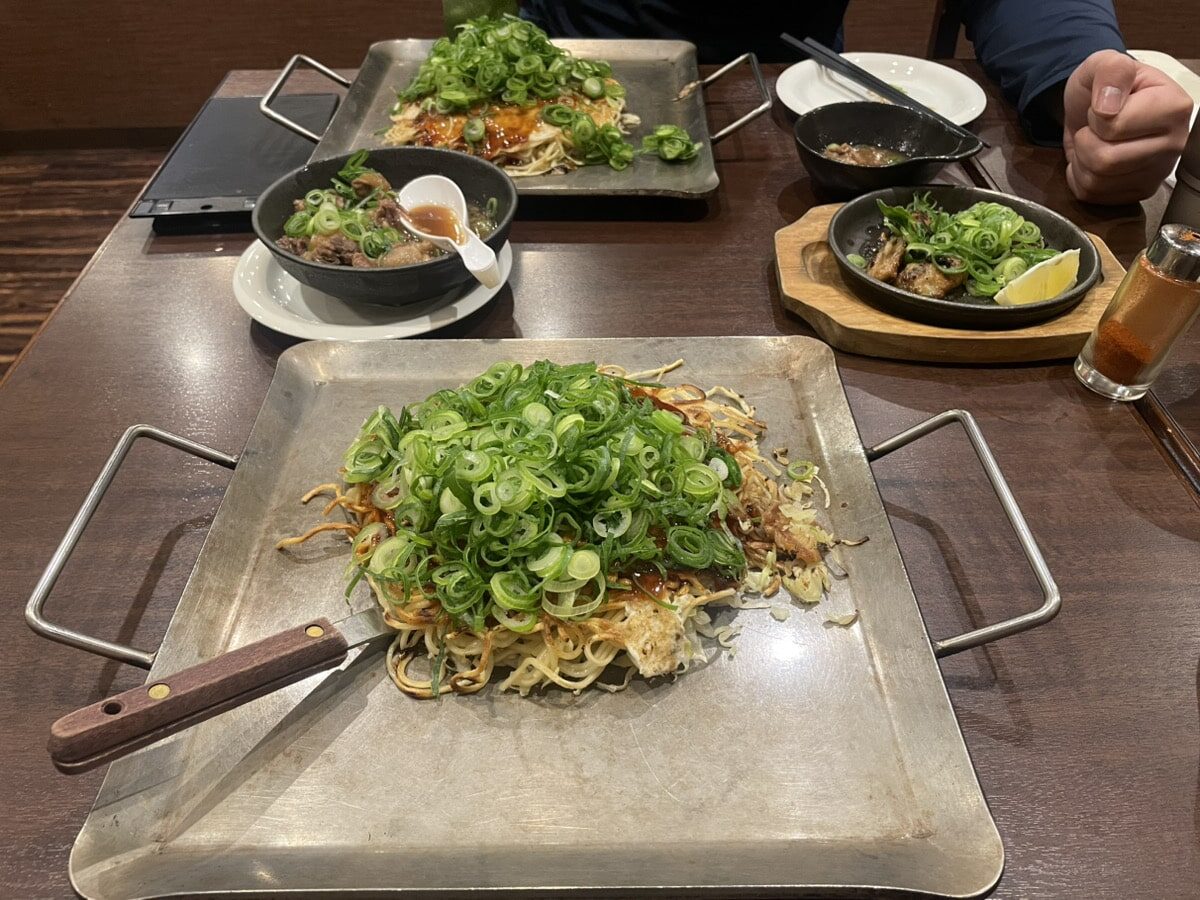 Two large metal trays of noodles topped with chopped green onions on a restaurant table, with side dishes, utensils, and drinks visible.