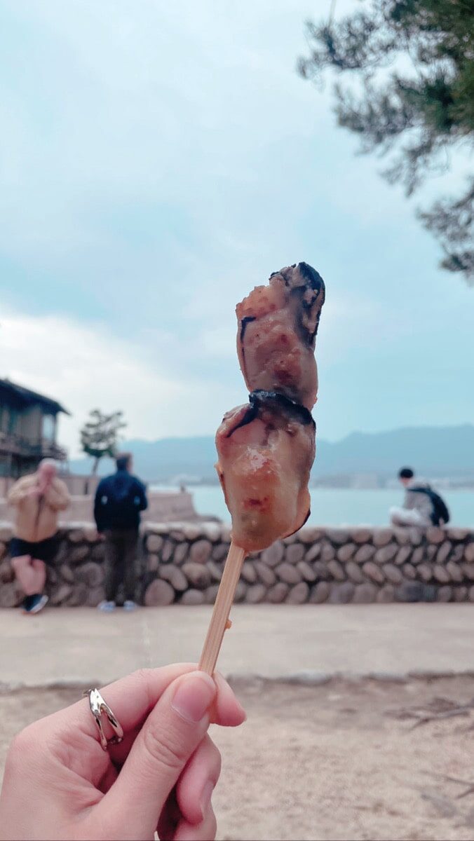 A hand holding a skewer with grilled seafood, outdoors near a stone wall with people, trees, and mountains in the background.