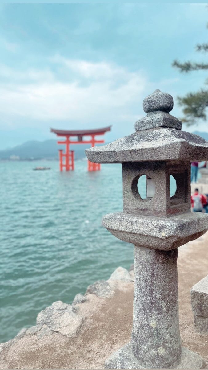 Stone lantern in the foreground with a red torii gate standing in the water in the background under a cloudy sky.