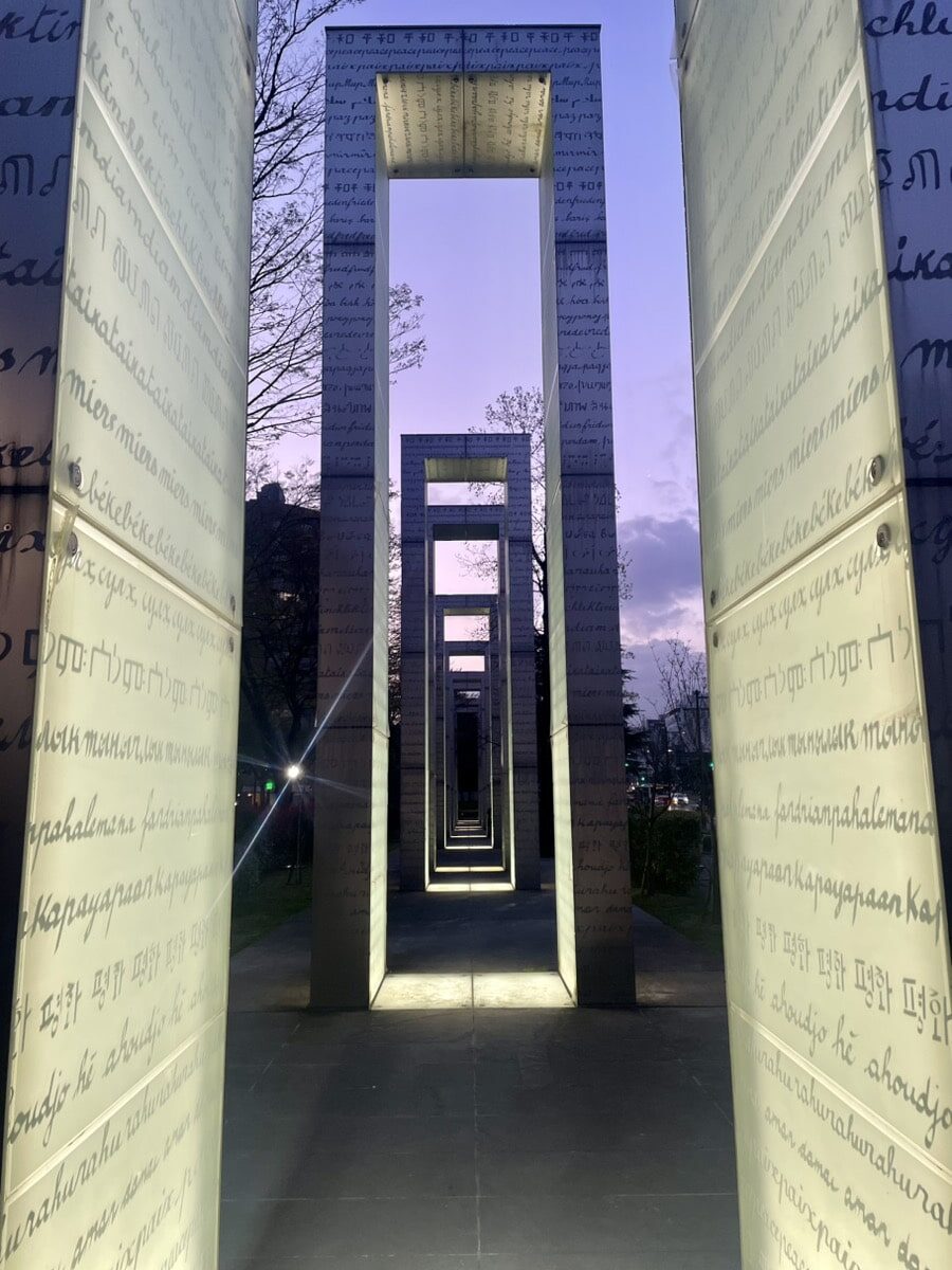 Rectangular metal monuments with engraved writing stand in a row, illuminated from below, with trees and a sunset sky in the background.