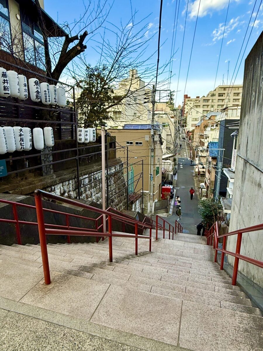 Stone staircase with red railings leading down to a narrow street lined with buildings; paper lanterns and trees are visible on the left.