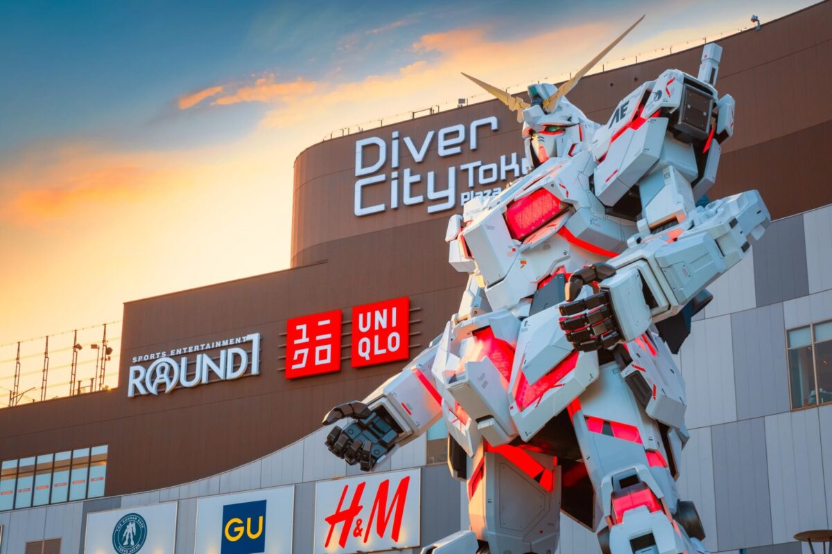 A large Gundam robot statue stands in front of DiverCity Tokyo Plaza shopping mall with store signs visible, including UNIQLO, GU, and H&M, at sunset.