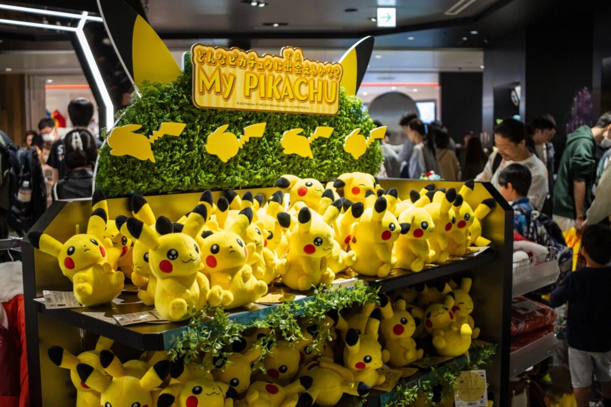 A store display filled with multiple Pikachu plush toys, with a sign reading "My Pikachu," and people browsing in the background.