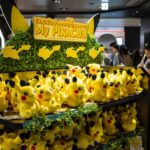 A store display filled with multiple Pikachu plush toys, with a sign reading "My Pikachu," and people browsing in the background.
