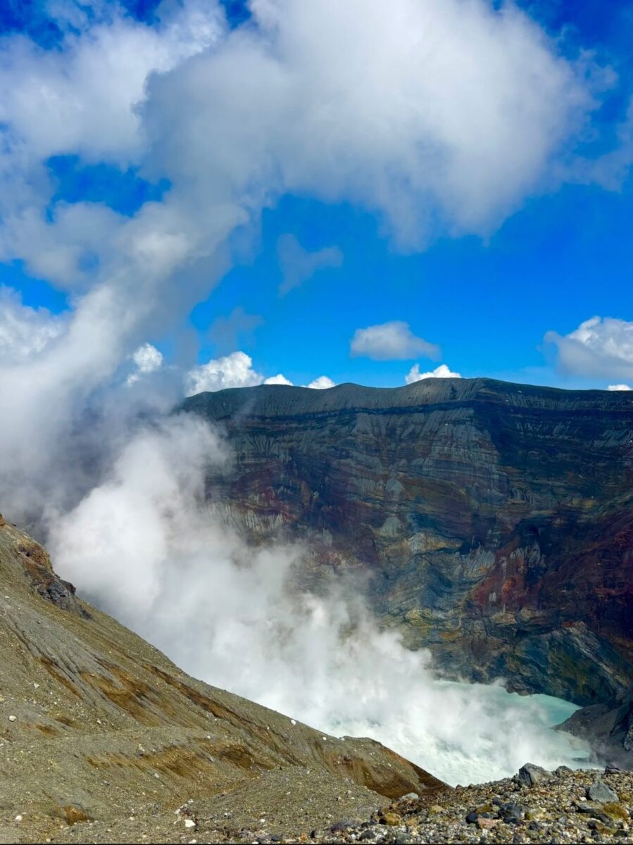 Steam rises from the crater of an active volcano under a bright blue sky, with rocky terrain and layered crater walls visible.