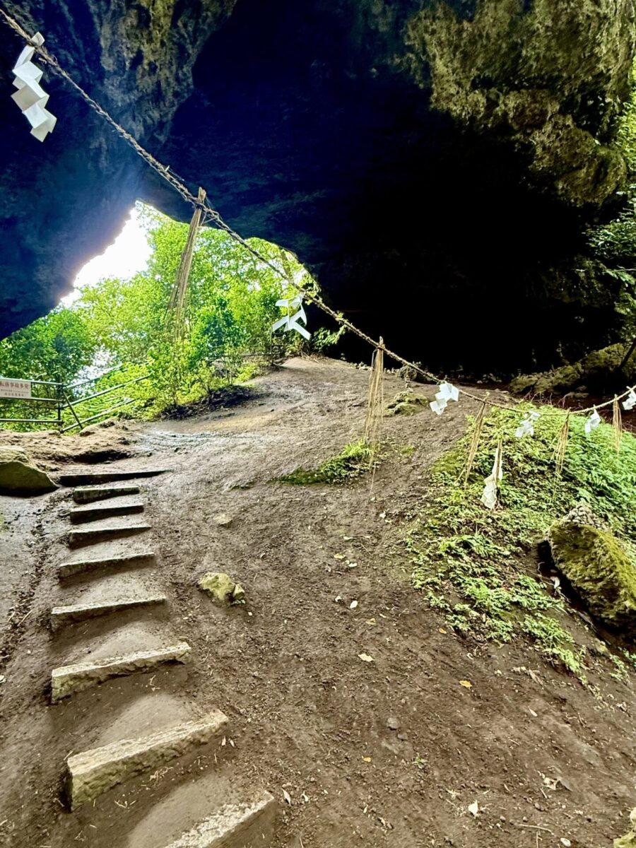 Stone steps lead into a cave entrance adorned with shimenawa rope and shide paper, surrounded by greenery and dappled sunlight.