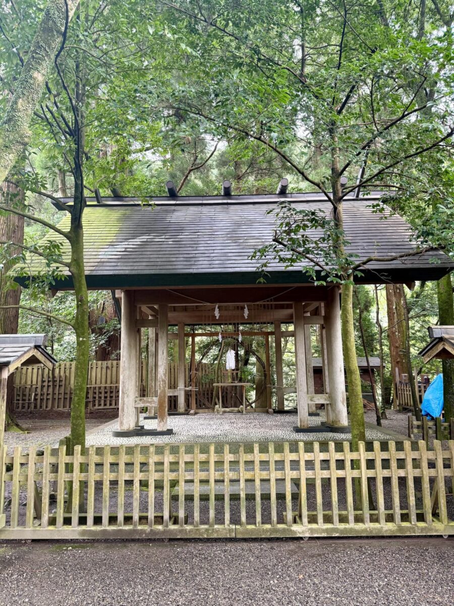 A traditional wooden Shinto shrine with a peaked roof stands in a forested area, surrounded by a wooden fence and trees.