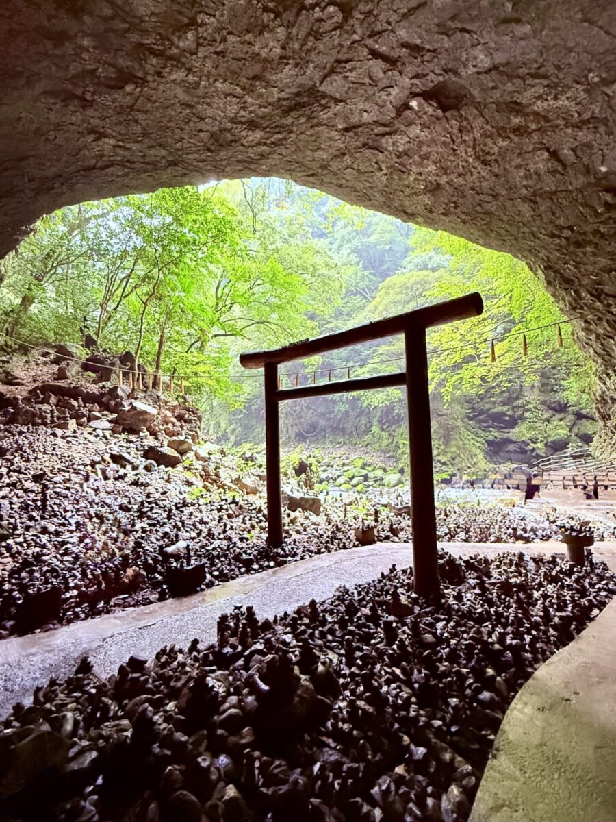 View from inside a cave looking out at a traditional Japanese torii gate, surrounded by small stone piles and lush green trees.