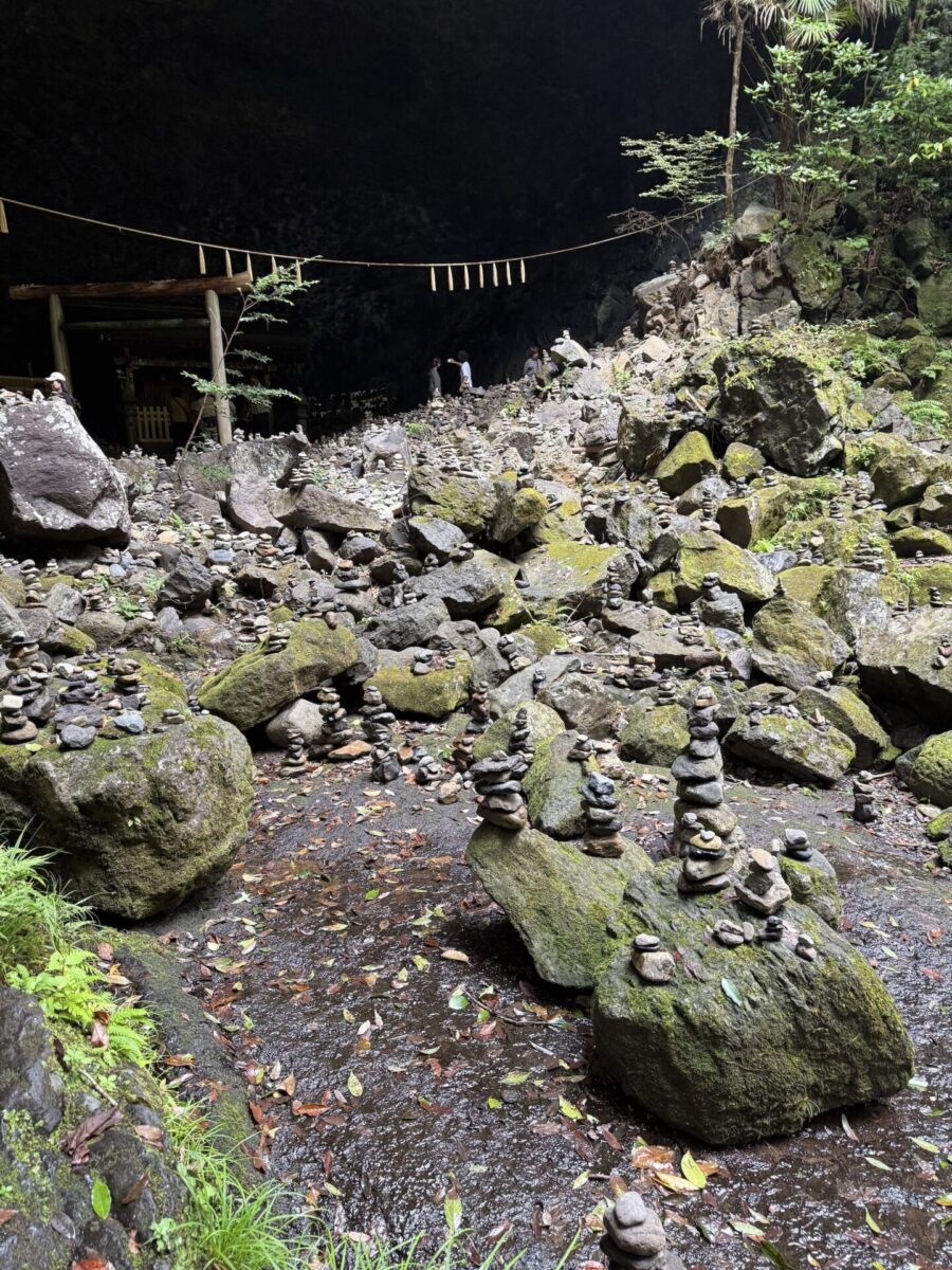 Stacks of small stones are arranged on mossy rocks at the entrance of a cave, with a wooden structure and hanging rope visible in the background.