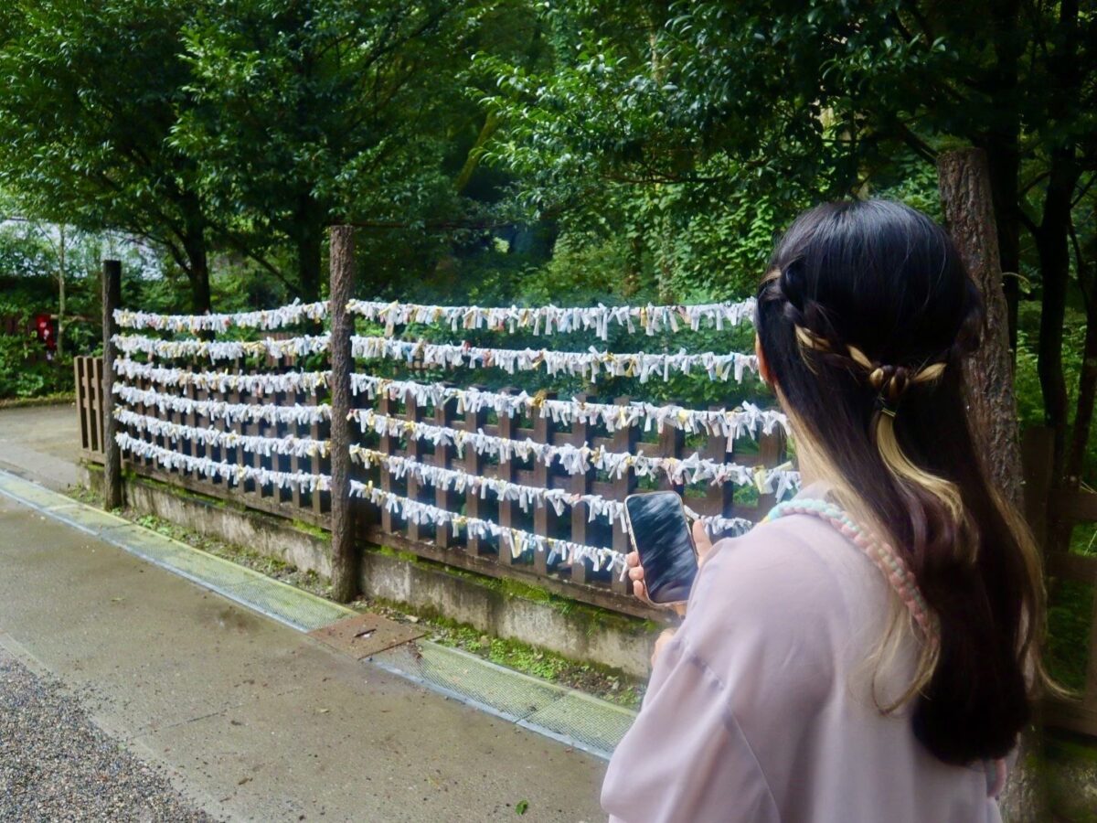 A woman holding a smartphone stands in front of wooden racks displaying numerous tied paper fortunes at a shrine, surrounded by trees.