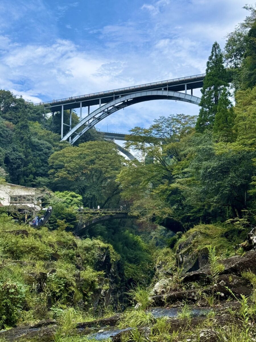 Three bridges, 2 modern and one older, span a lush green gorge with trees, vegetation, and a rocky stream below under a partly cloudy sky.