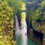 A narrow river runs between tall, green cliffs with a waterfall cascading into the water. Several people are boating below, and a bridge spans the gorge above.