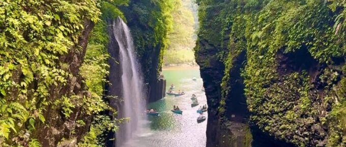 A narrow river runs between tall, green cliffs with a waterfall cascading into the water. Several people are boating below, and a bridge spans the gorge above.