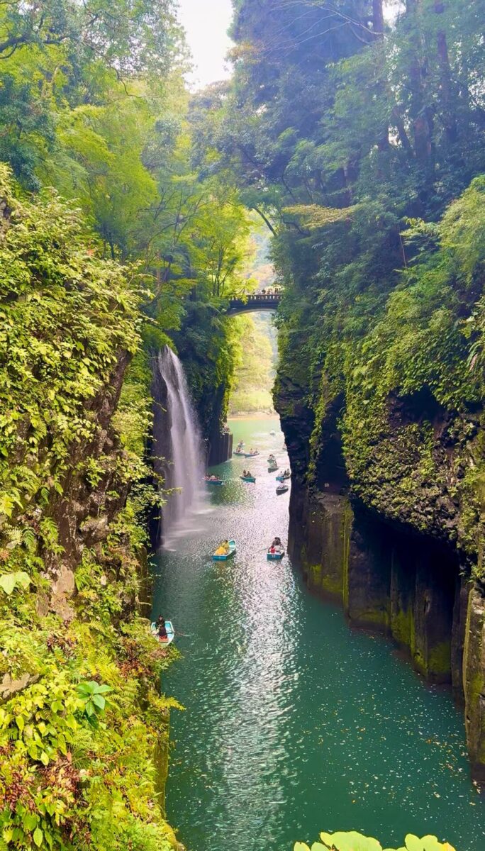 A narrow river runs between tall, green cliffs with a waterfall cascading into the water. Several people are boating below, and a bridge spans the gorge above.